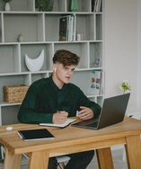 Young man writing notes at his desk next to a laptop