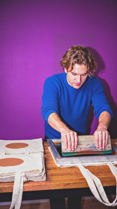 A young man prints his own tote bags at a table at home.